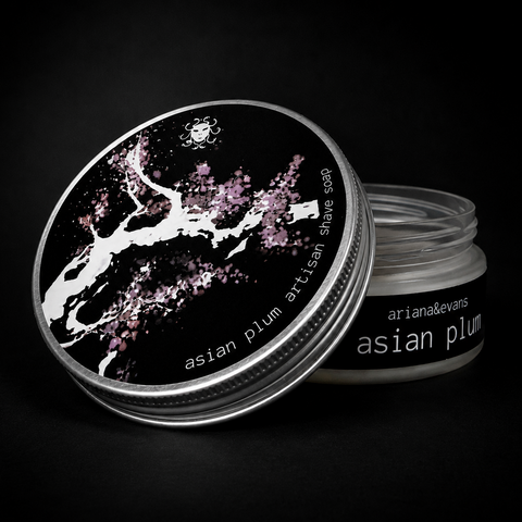 A silver metal jar of Asian Plum Artisan Shave soap with its lid leaning against it, set against a solid black background. The lid features a black label with a white cherry blossom tree design and pink plum blossoms.